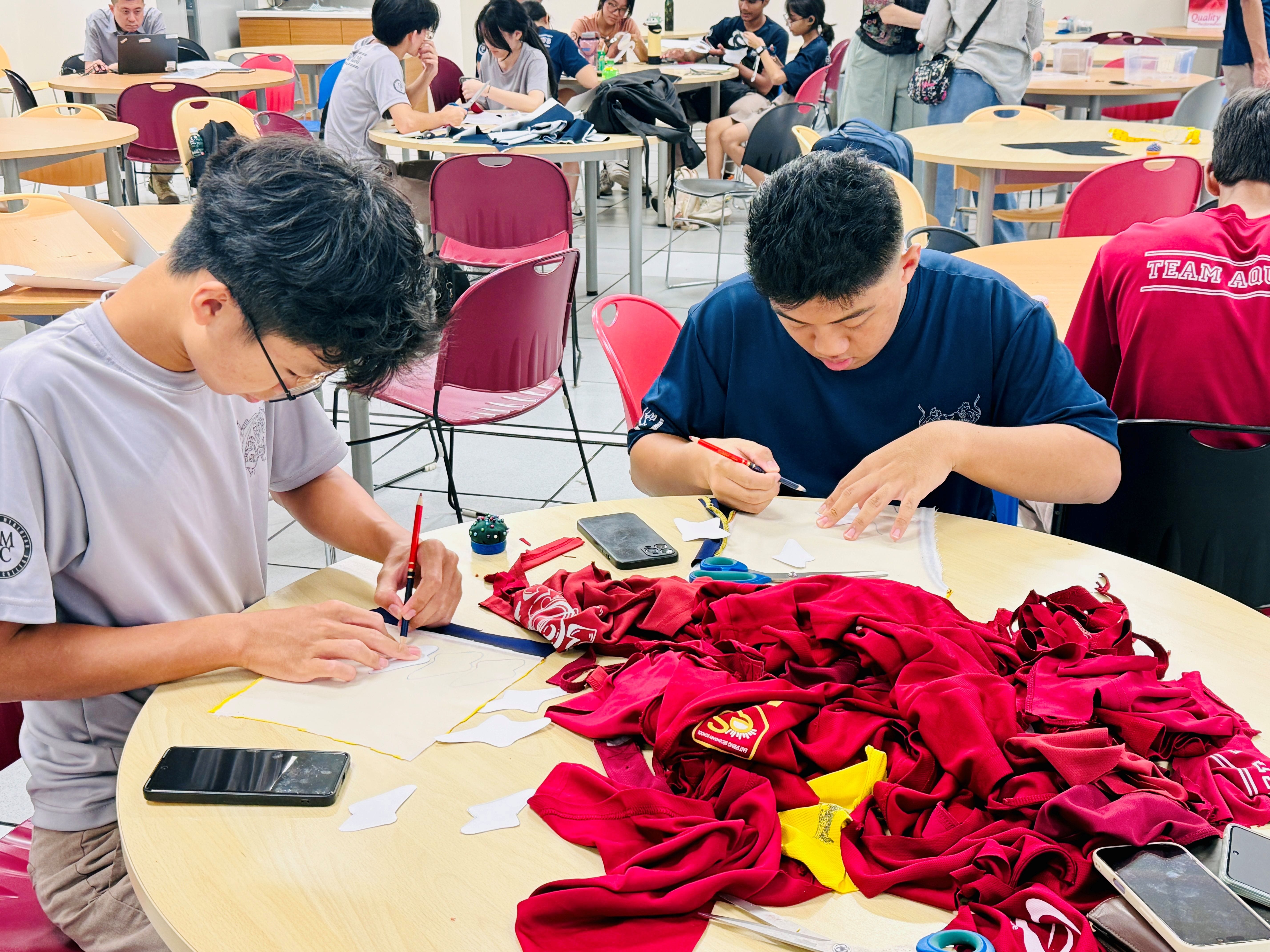 Two male students sit at a round table covered in red fabric and clothing, carefully tracing or drawing patterns onto white fabric pieces using red markers, with other students working in the background.